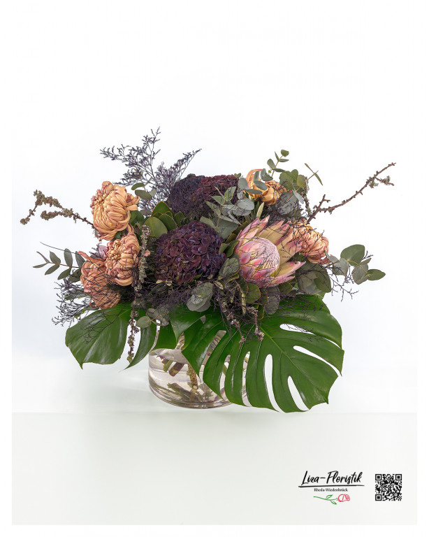 Ein prachtvoller Blumenstrauß mit exotischer Königsprotea, dekorativen Lärchenzweigen, edlen Hortensien, zarten Chrysanthemen, frischem Eukalyptus, filigraner Statice und imposanten Monsterablättern – eine harmonische Kombination aus natürlichen Farben und Strukturen, die Eleganz und Exotik vereint.