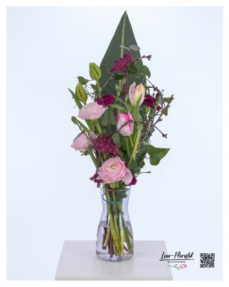 Moderner Blumenstrauß mit rosa Ranunkeln, violetten Nelken, gefärbtem Ammi und zarten Tulpen, kombiniert mit dekorativen Kirschzweigen und einem großen Strelitzienblatt als Hintergrund.